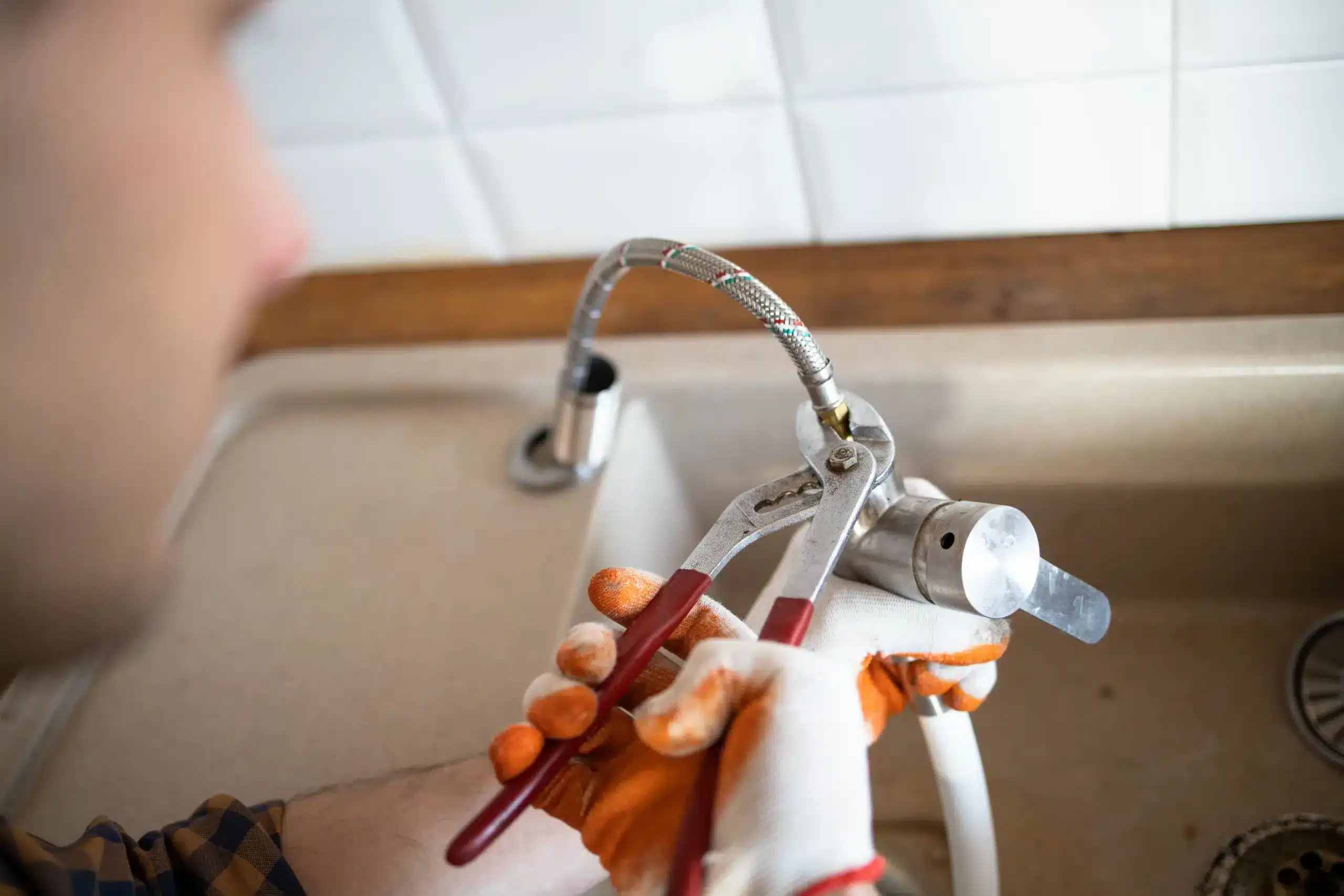 Gloved plumber using pliers to fix a kitchen faucet with a braided hose